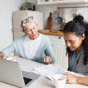 Black girl volunteer or social worker helping elderly lonely woman to deal with documents, agreement or invoice, reading and examining paper doc sitting at kitchen table together in front of laptop
